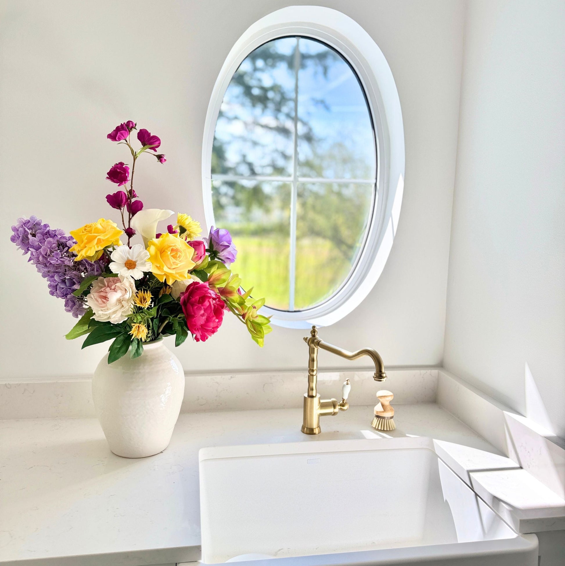 Bathroom with a white sink, colorful and realistic artificial flowers in a vase, and a round window. Faux peonies, faux roses, faux hydrangea, and faux orchids in an artificial flower bouquet for My Faux Florist Canada.