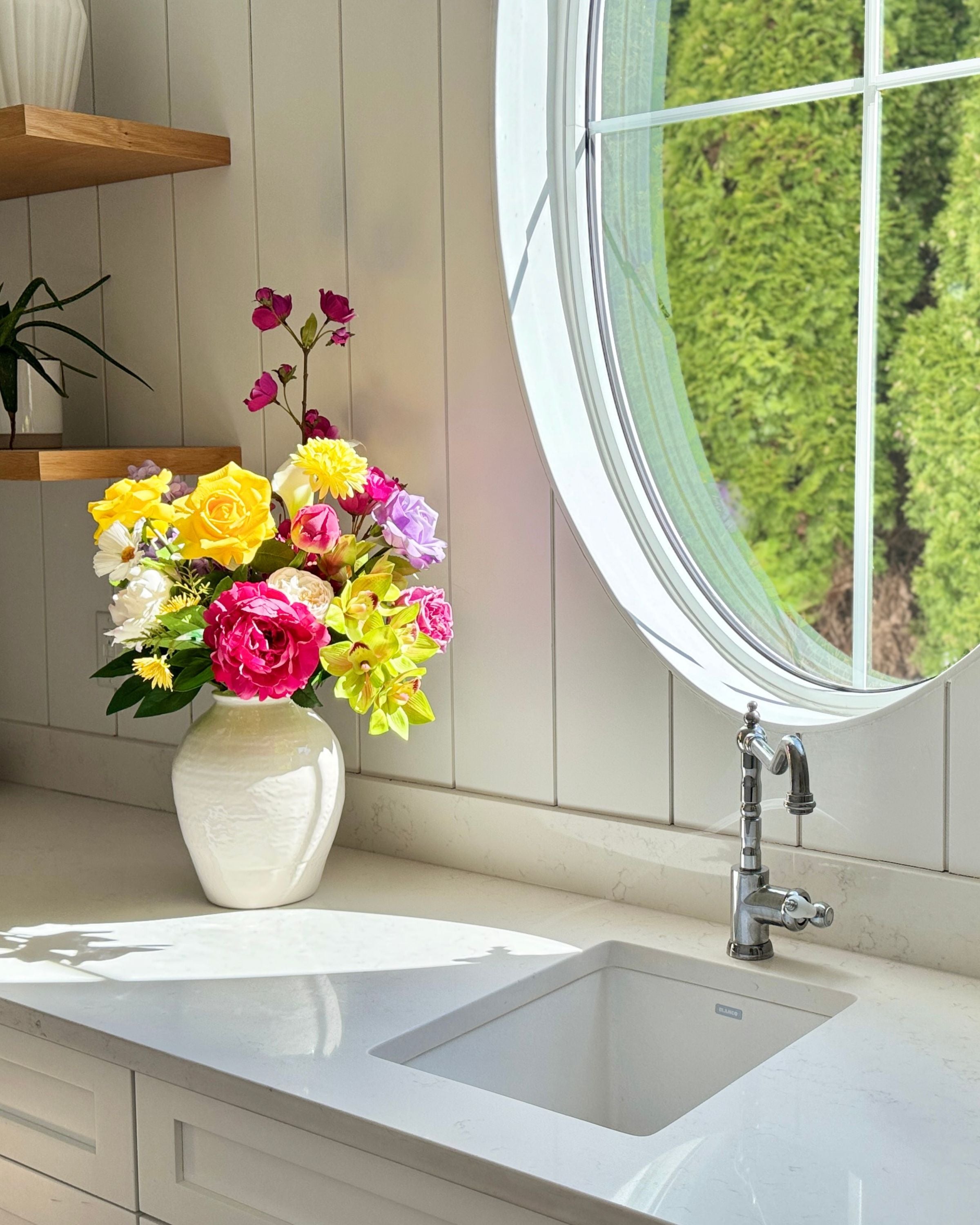Colorful and realistic artificial flowers in a vase on a kitchen counter with a round window in the background. Faux peonies, faux roses, faux hydrangea, and faux orchids in an artificial flower bouquet for My Faux Florist Canada.