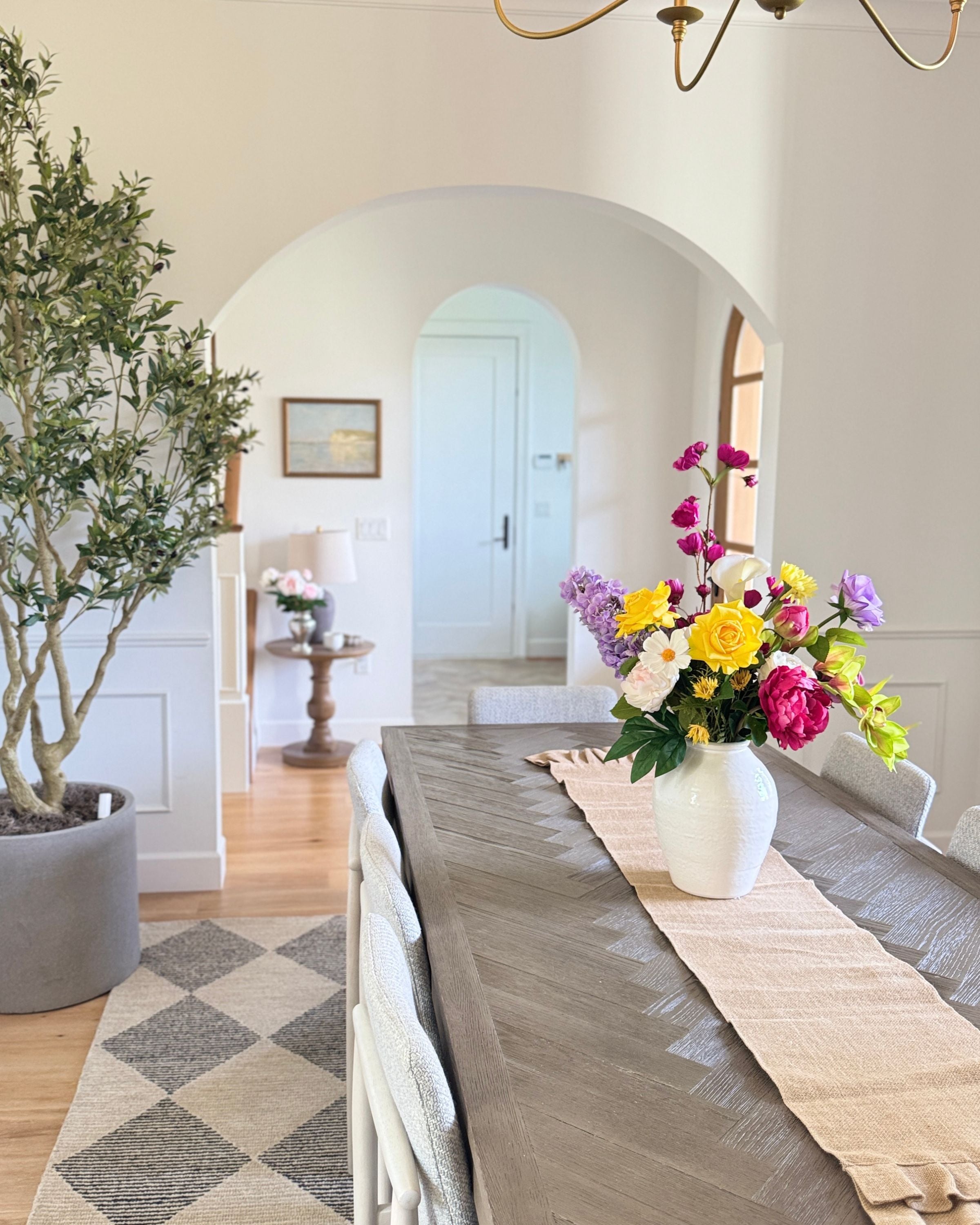 Dining room with a long table set for a meal, realistic artificial flowers in a vase, and a large mirror on the wall. Faux peonies, faux roses, faux hydrangea, and faux orchids in an artificial flower bouquet for My Faux Florist Canada.