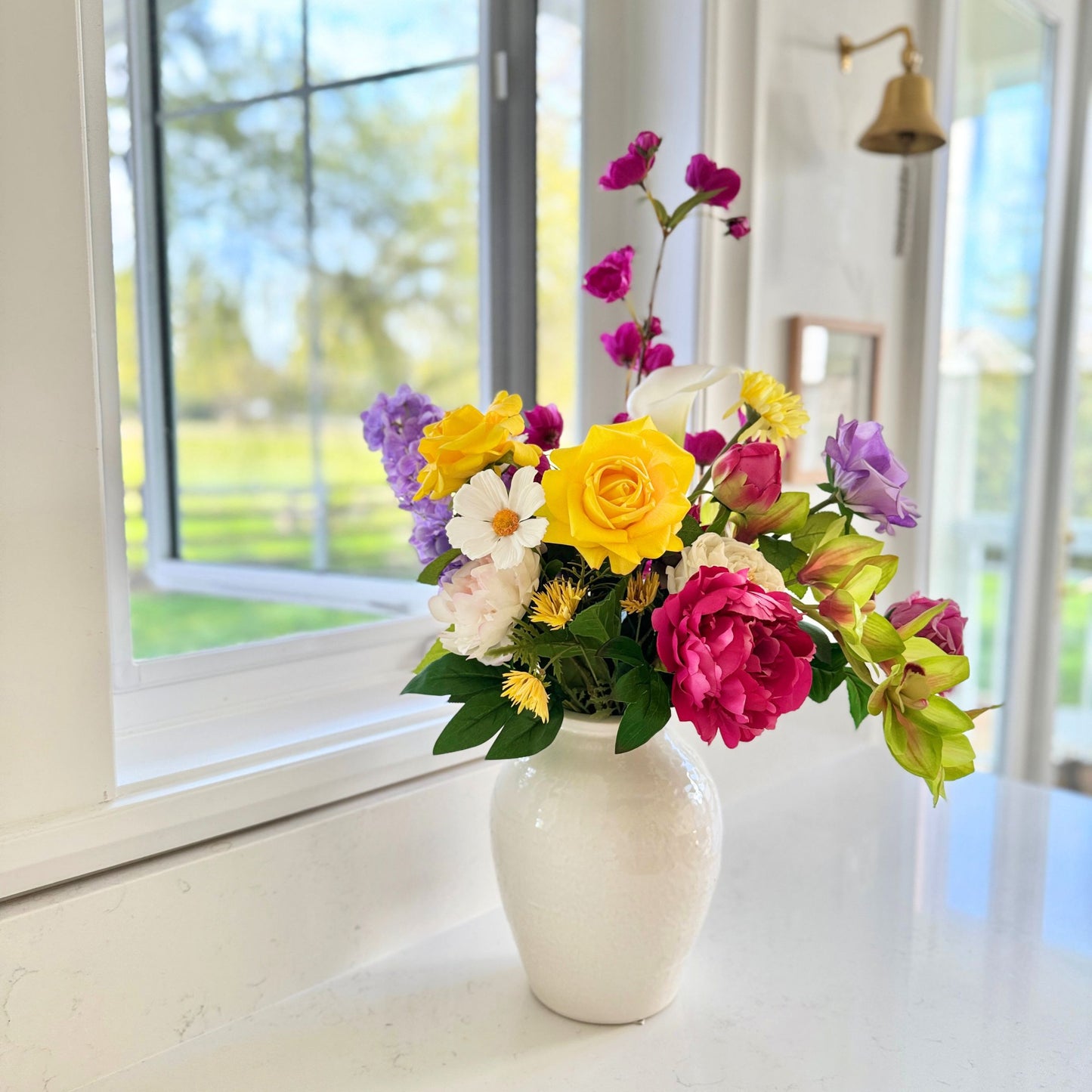 Colorful and realistic artificial flower arrangement in a white vase on a windowsill with a view of greenery outside. Faux peonies, faux roses, faux hydrangea, and faux orchids in an artificial flower bouquet for My Faux Florist Canada.