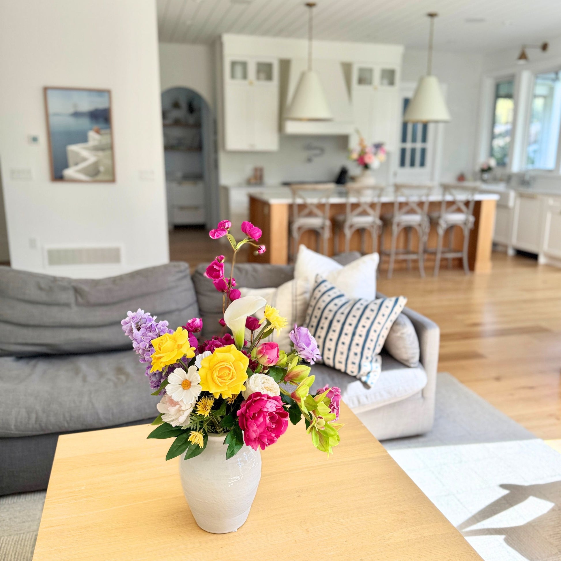 Colorful and realistic artificial flowers in a vase on a coffee table with a living room and kitchen in the background. Faux peonies, faux roses, faux hydrangea, and faux orchids in an artificial flower bouquet for My Faux Florist Canada.