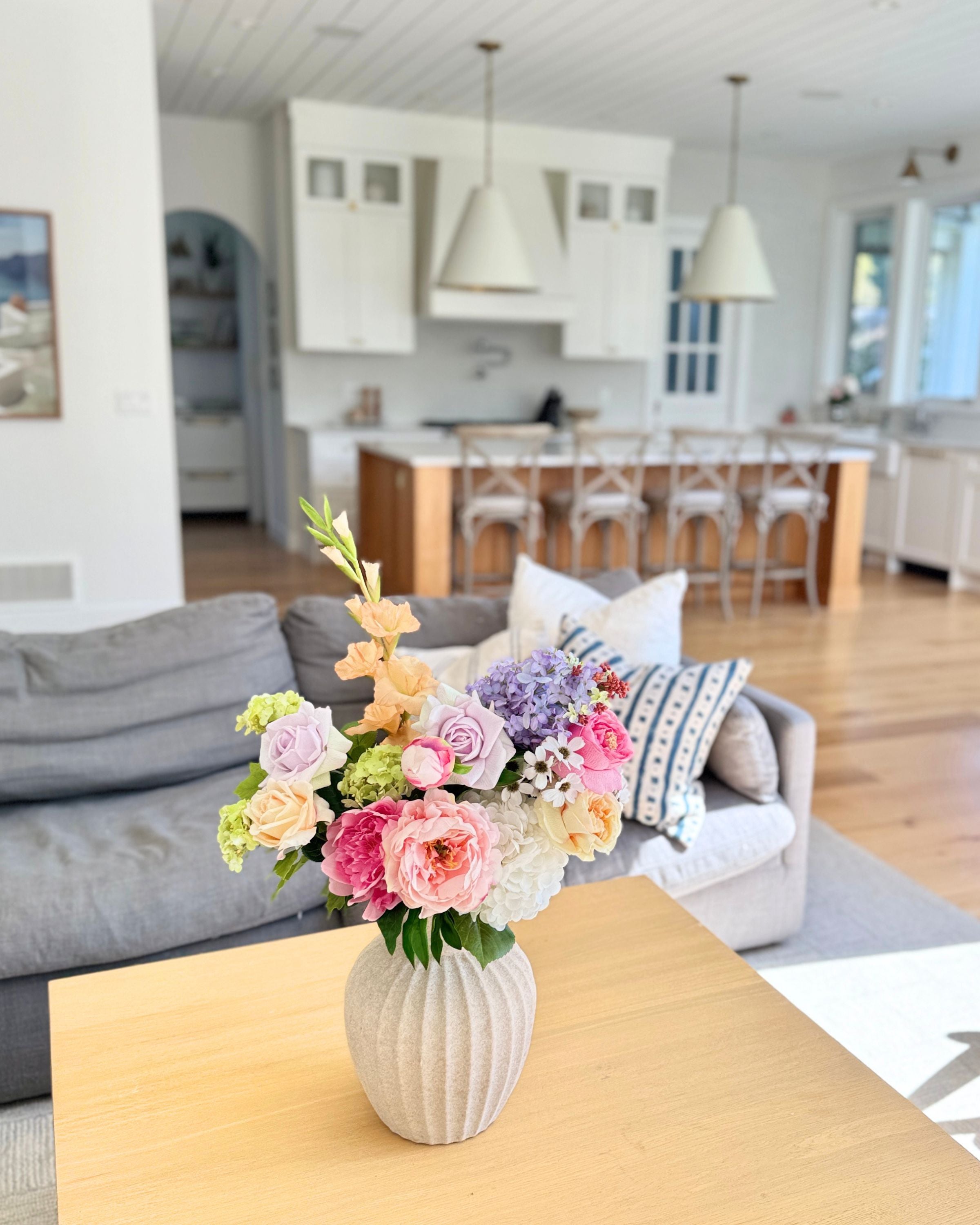 A vase of colourful, realistic artificial flowers sits on a living room table with a kitchen in the background. Faux peonies, faux roses, faux hydrangea, and faux orchids in an artificial flower bouquet for My Faux Florist Canada.