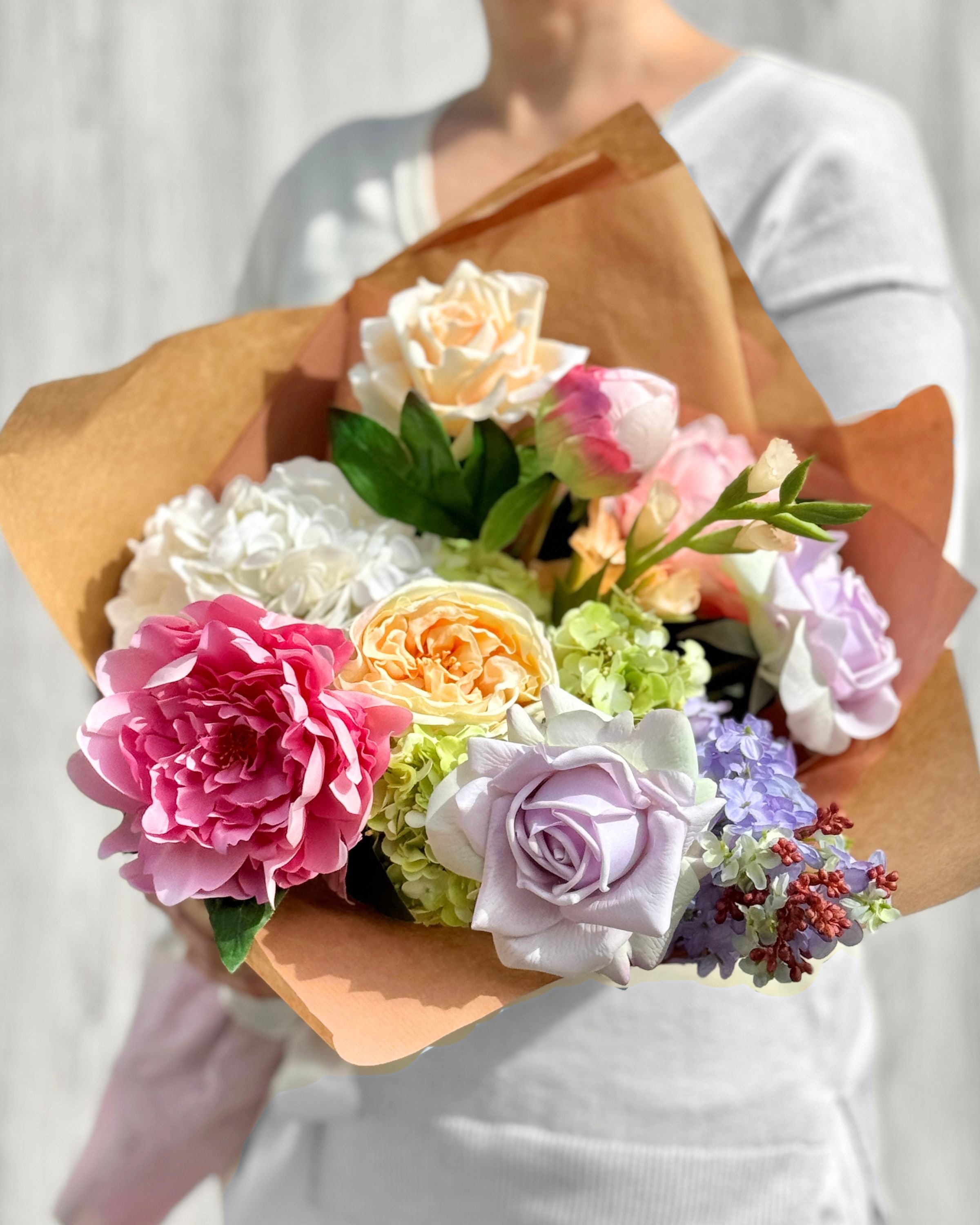 Bouquet of colorful and realistic artificial flowers wrapped in brown paper held by a person. Faux peonies, faux roses, faux hydrangea, and faux orchids in an artificial flower bouquet for My Faux Florist Canada.