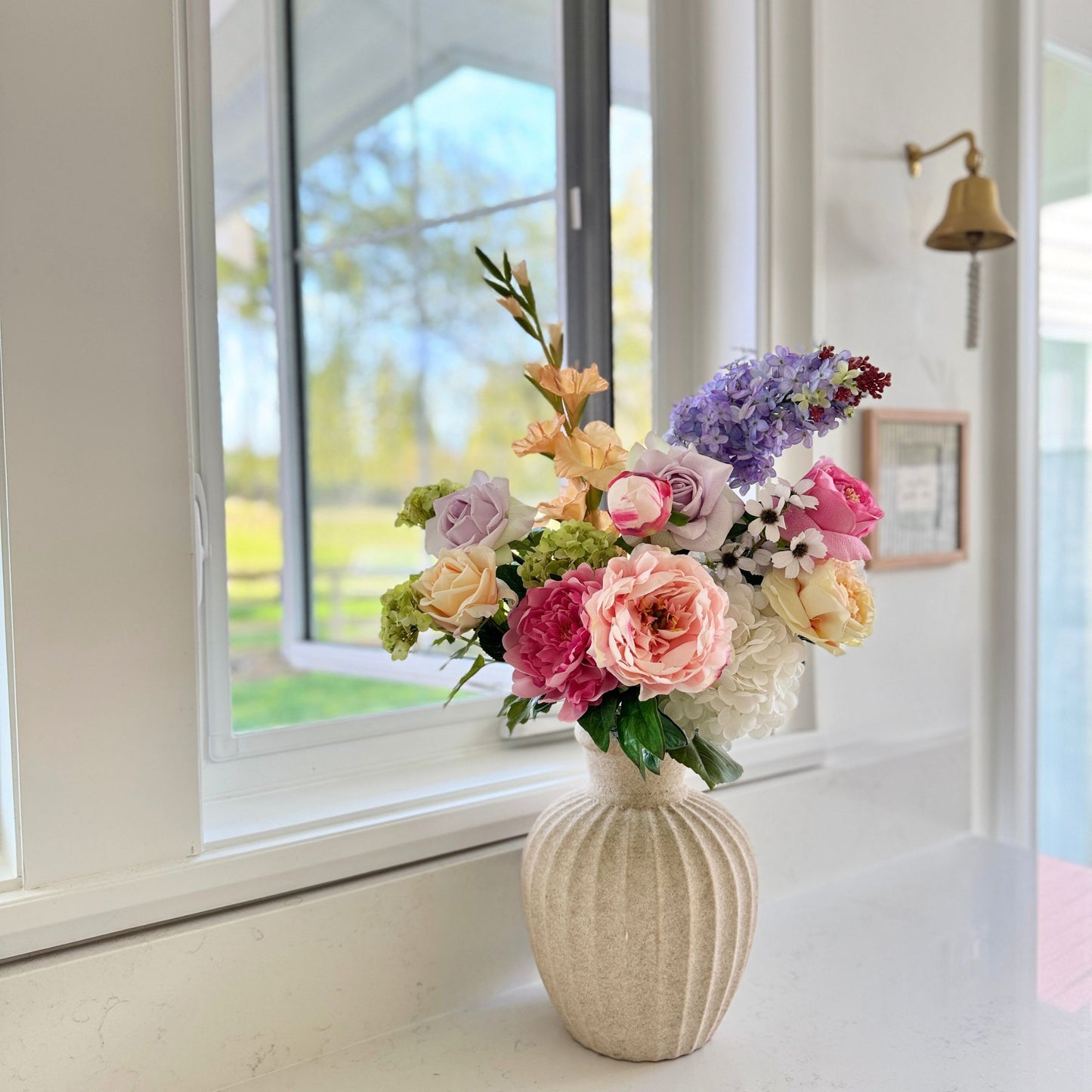 Bouquet of realistic artificial flowers in a woven vase on a windowsill with a view of greenery outside. Faux peonies, faux roses, faux hydrangea  for My Faux Florist Canada.