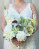 Bride holding a realistic artificial flower bridal bouquet featuring pale green artificial hydrangeas, white artificial dahlias, white artificial peonies, white artificial tea roses, white artificial Austin roses, and deep wine artificial calla lilies accented with soft lamb’s ear greenery — from My Faux Florist Canada.
