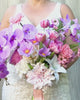 Bride holding a realistic artificial flower bridal bouquet featuring purple artificial orchids, pink artificial cosmos, champagne artificial tea roses, blush artificial peonies, white artificial hydrangeas, mauve artificial dahlias, and pink artificial tulips — from My Faux Florist Canada.
