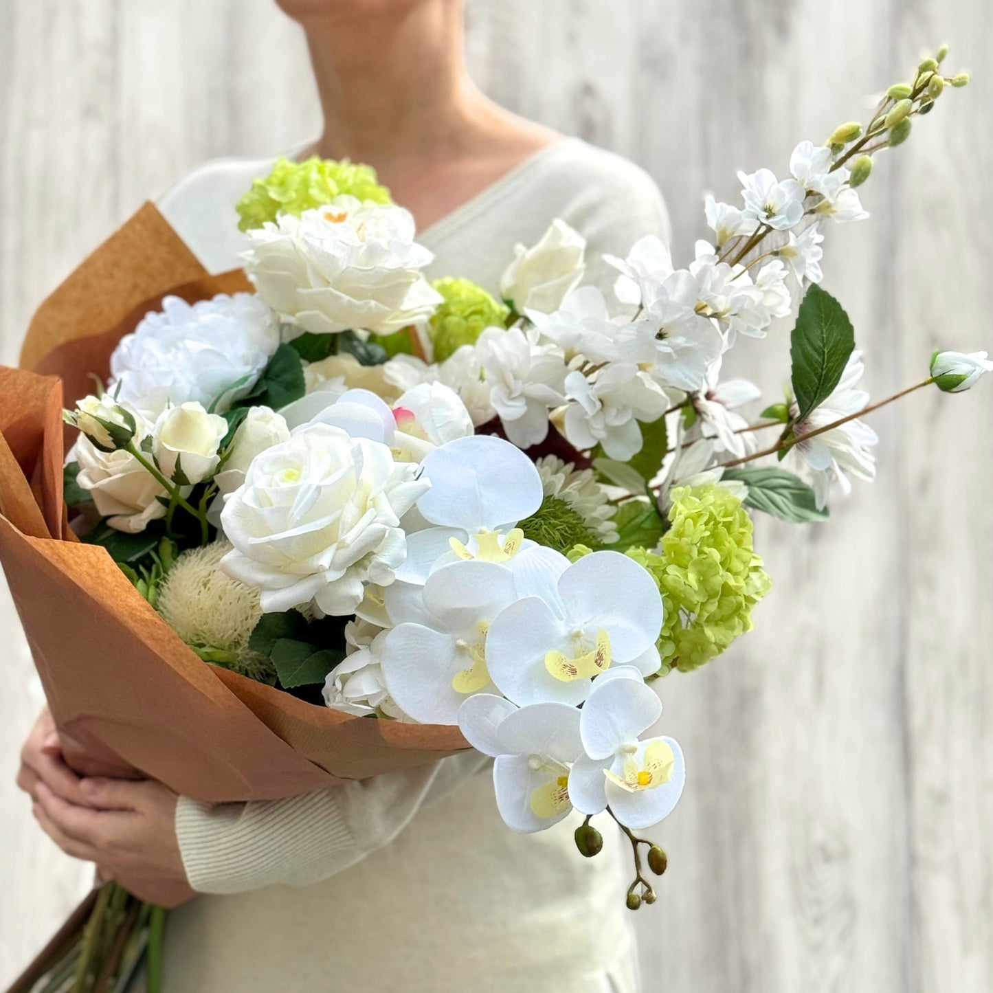 Bouquet of white and green realistic artificial flowers held by a person against a neutral background. Faux peonies, faux roses, faux hydrangea, and faux orchids in an artificial flower bouquet for My Faux Florist Canada.