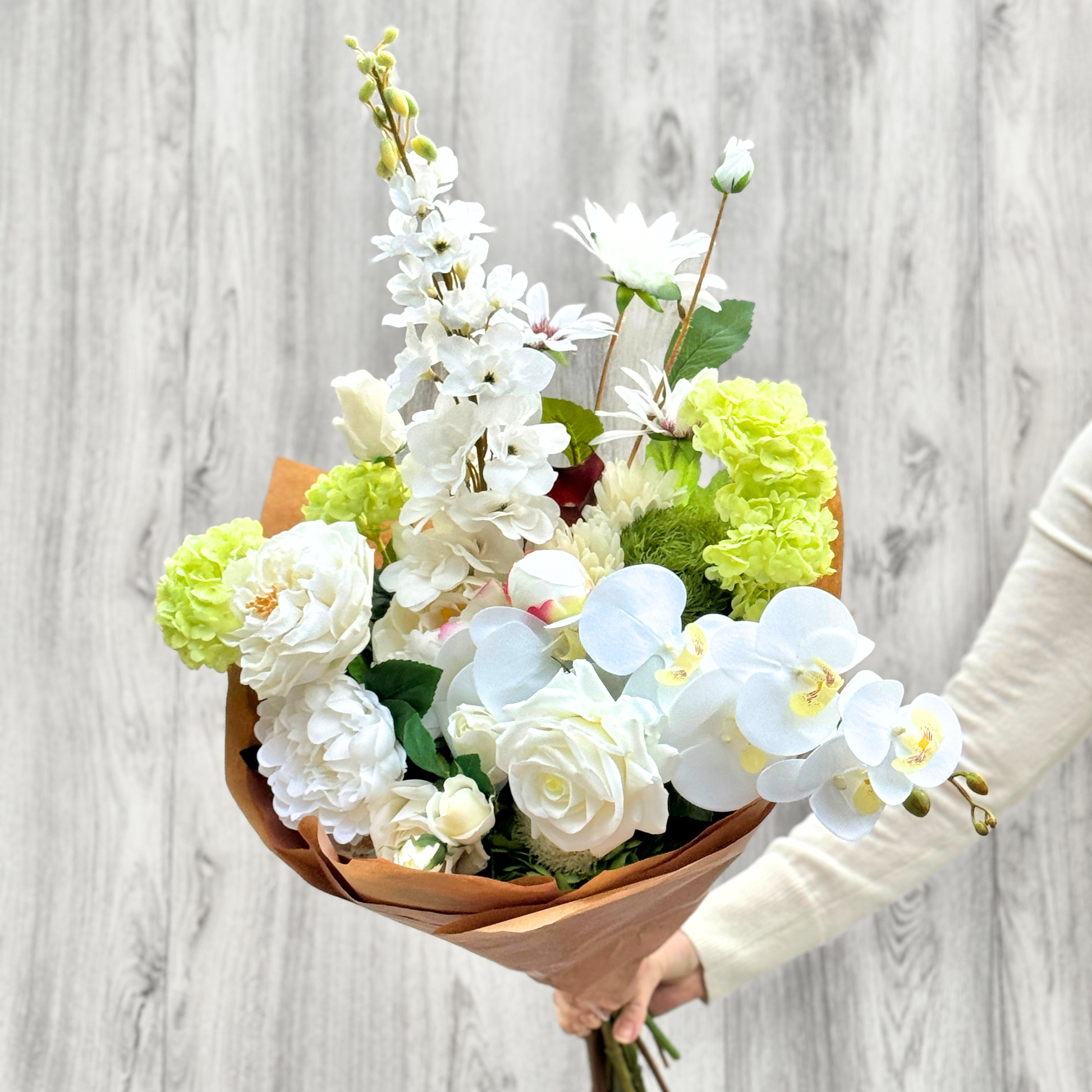 Bouquet of white and green realistic artificial flowers held by a person against a light gray wooden background. Faux peonies, faux roses, faux hydrangea, and faux orchids in an artificial flower bouquet for My Faux Florist Canada.