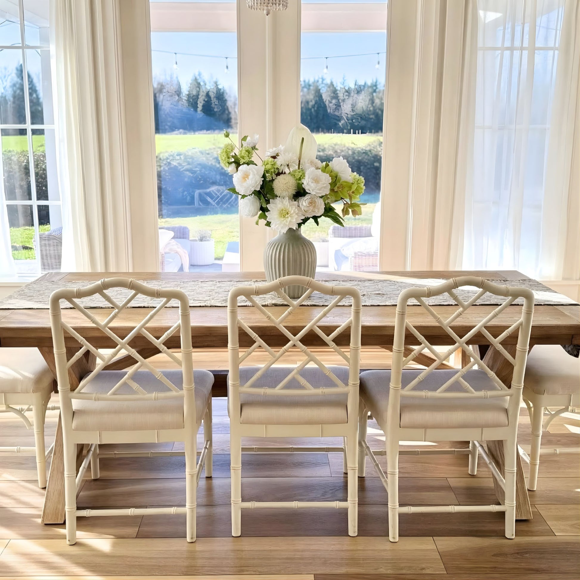 Dining room with wooden table and white chairs, large windows with curtains, and a vase of realistic artificial flowers. Faux peonies, faux roses, faux hydrangea, and faux orchids in an artificial flower bouquet for My Faux Florist Canada.