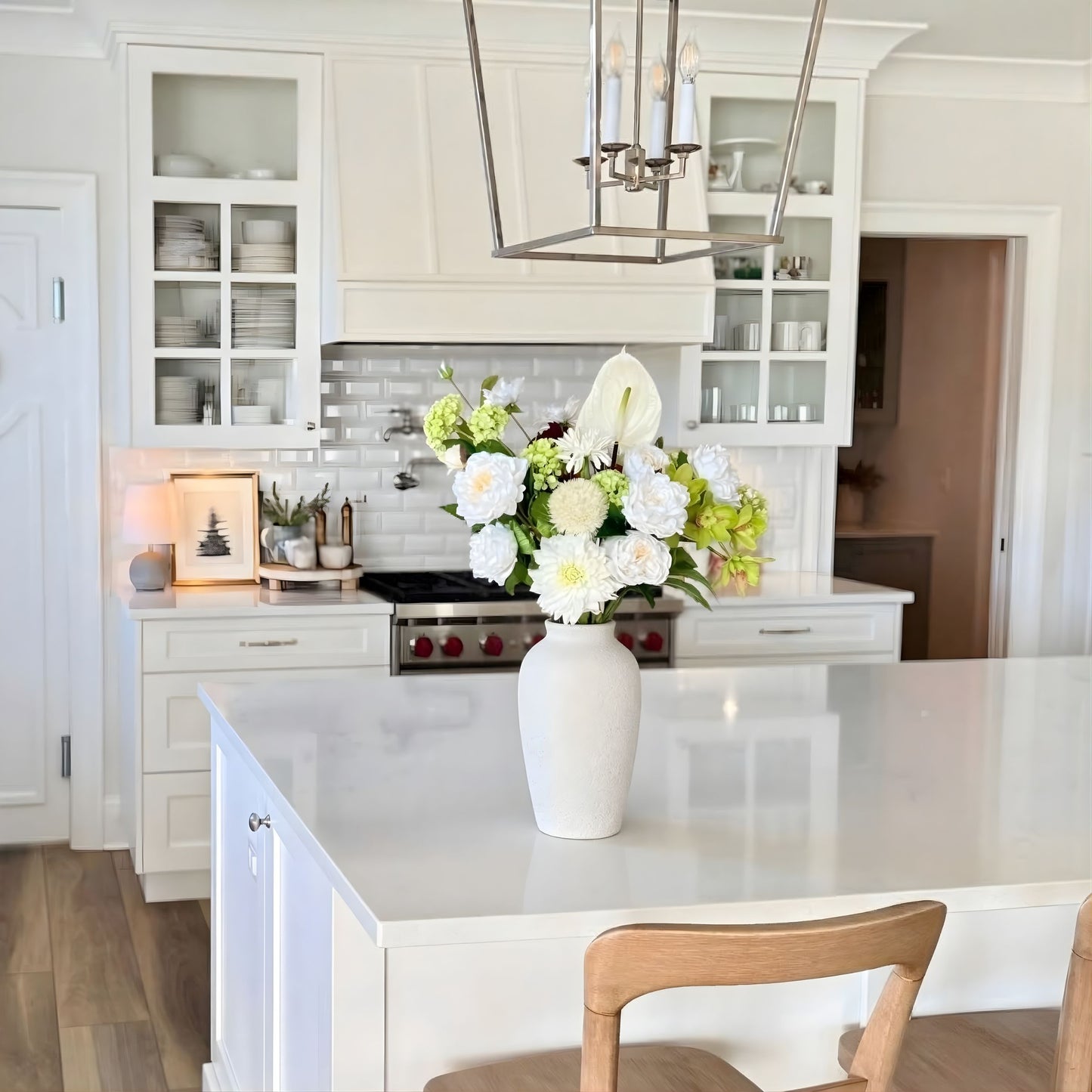 Modern kitchen with white cabinets, a vase of realistic artificial flowers on the counter, and a chandelier. Faux peonies, faux roses, faux hydrangea, and faux orchids in an artificial flower bouquet for My Faux Florist Canada.