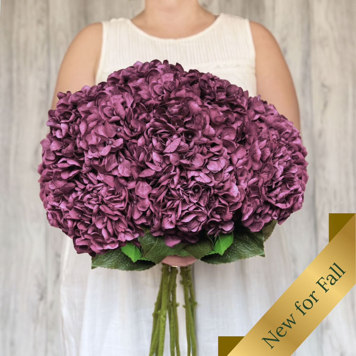 A person in a white sleeveless top holds a large round bouquet of deep purple realistic artificial hydrangea flowers with green stems and leaves, set against a light wood-paneled background. A gold diagonal banner in the lower right corner reads 'New for Fall'. From My Faux Florist Canada.