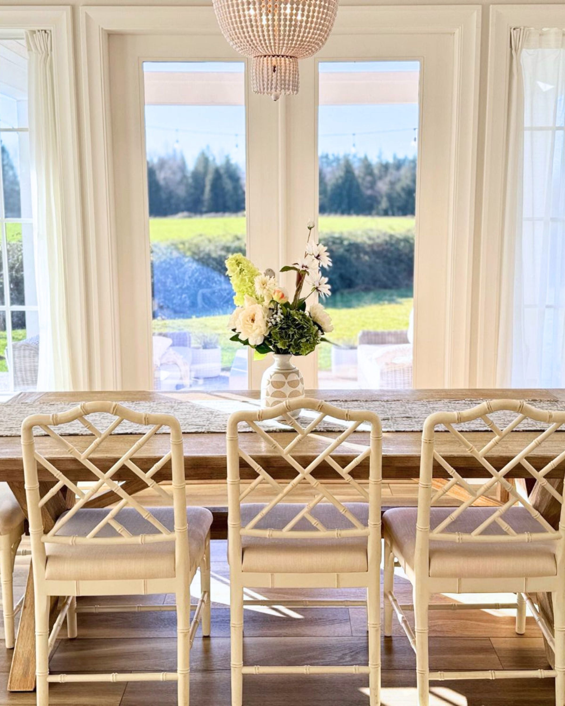Dining room with a long wooden table and chairs, a vase of realistic artificial flowers in the middle, and a large windows with scenic view. Faux peonies, faux roses, faux hydrangea, in an artificial flower bouquet for My Faux Florist Canada.