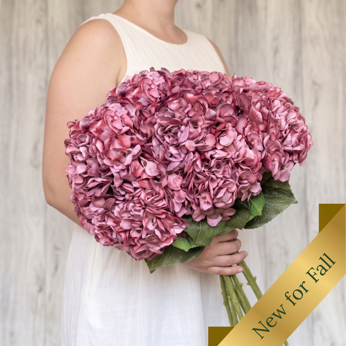 A person in a white sleeveless dress holds a large bouquet of pink realistic artificial hydrangea flowers with green leaves. A gold diagonal banner in the lower right corner reads 'New for Fall'. From My Faux Florist Canada.
