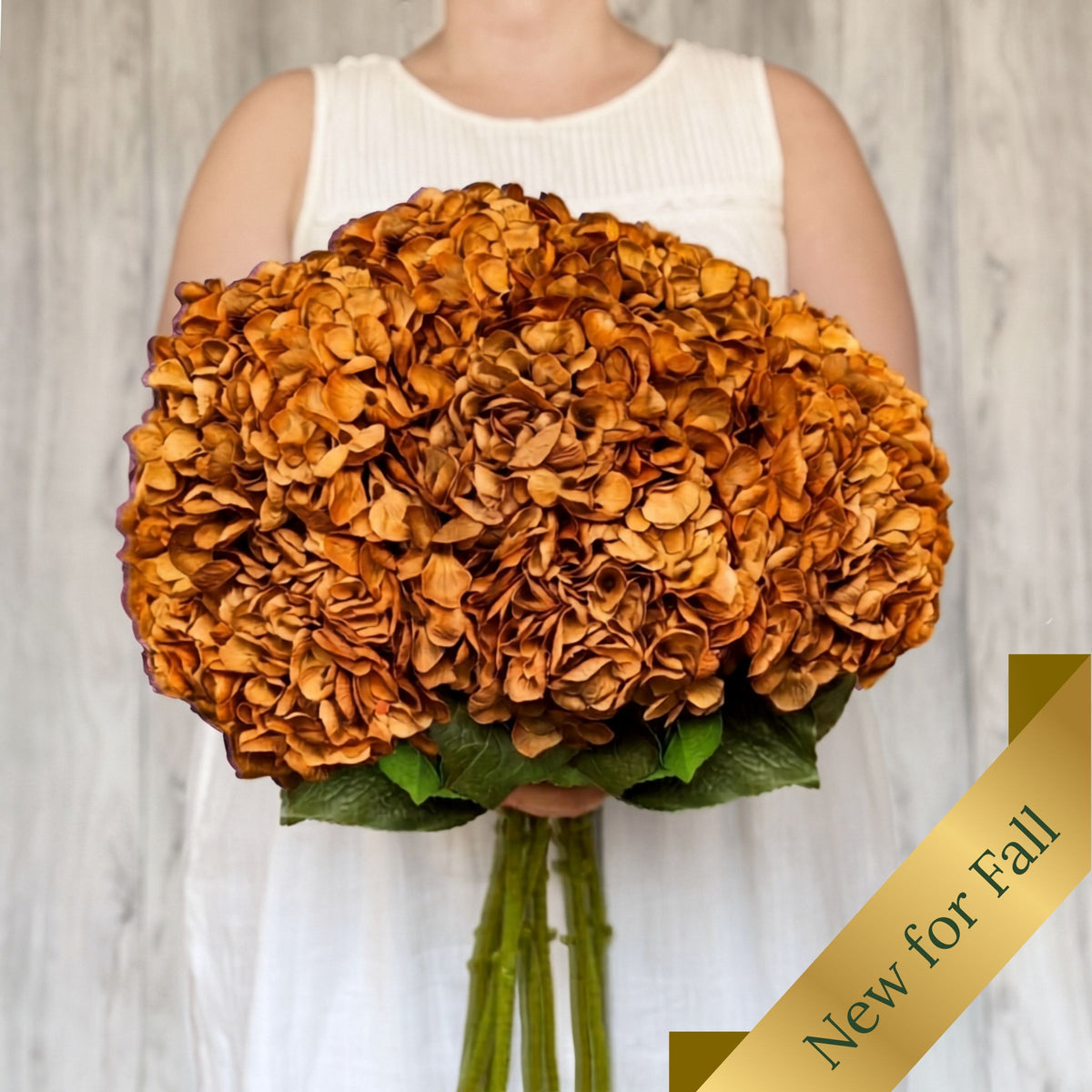 A person in a white sleeveless top holds a large round bouquet of realistic orange-brown faux hydrangea flowers with green stems against a light wood wall. A gold diagonal banner in the lower right reads 'New for Fall'. From My Faux Florist Canada.
