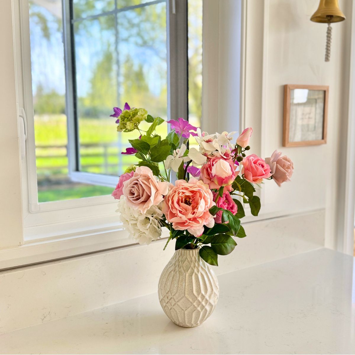 Vase with pink and white realistic artificial flowers on a windowsill with a view of greenery outside. Faux peonies, faux roses, faux hydrangea, in an artificial flower bouquet for My Faux Florist Canada.
