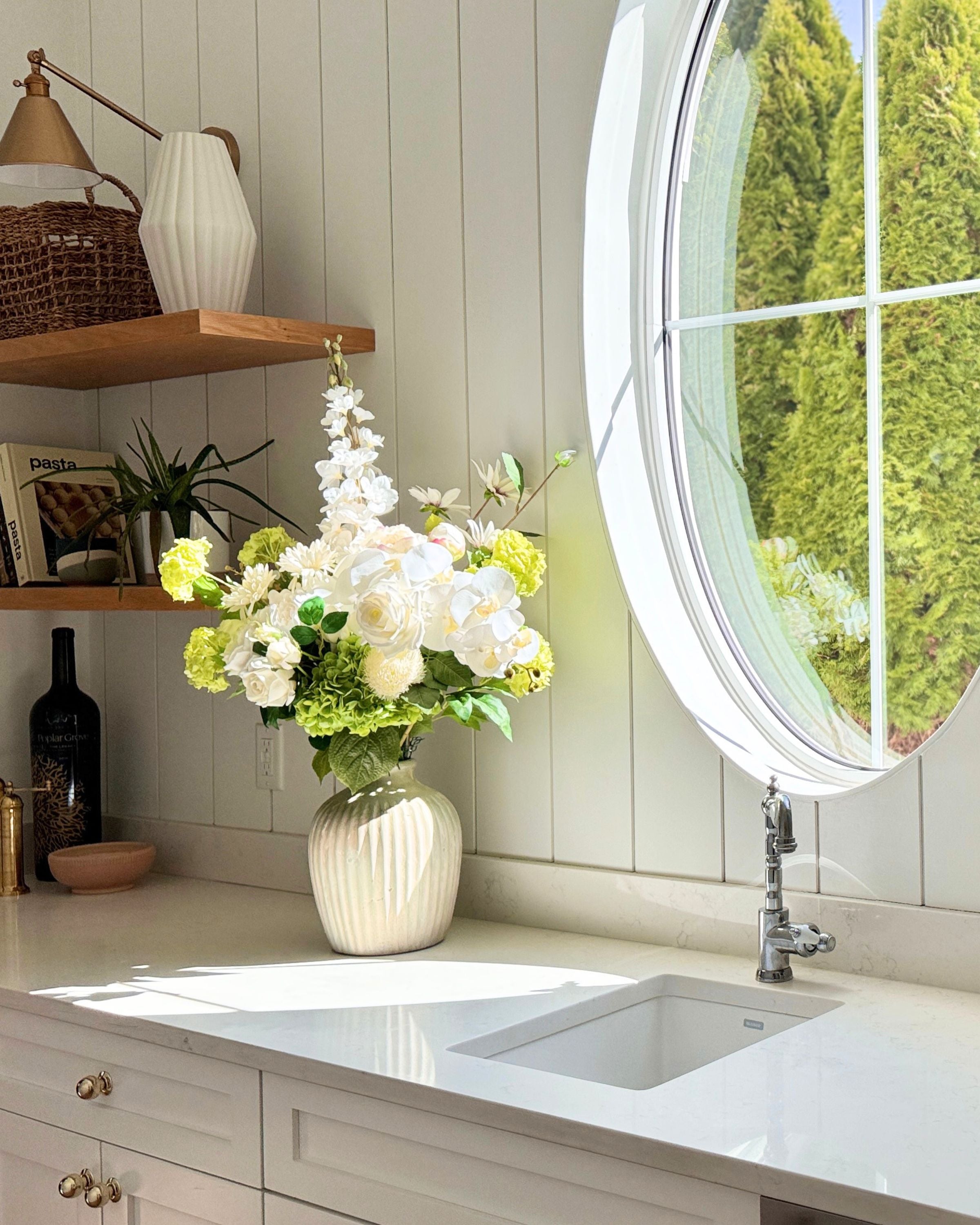 Kitchen counter with a vase of realistic artificial flowers, sink, and round window. Faux peonies, faux roses, faux hydrangea, and faux orchids in an artificial flower bouquet for My Faux Florist Canada.
