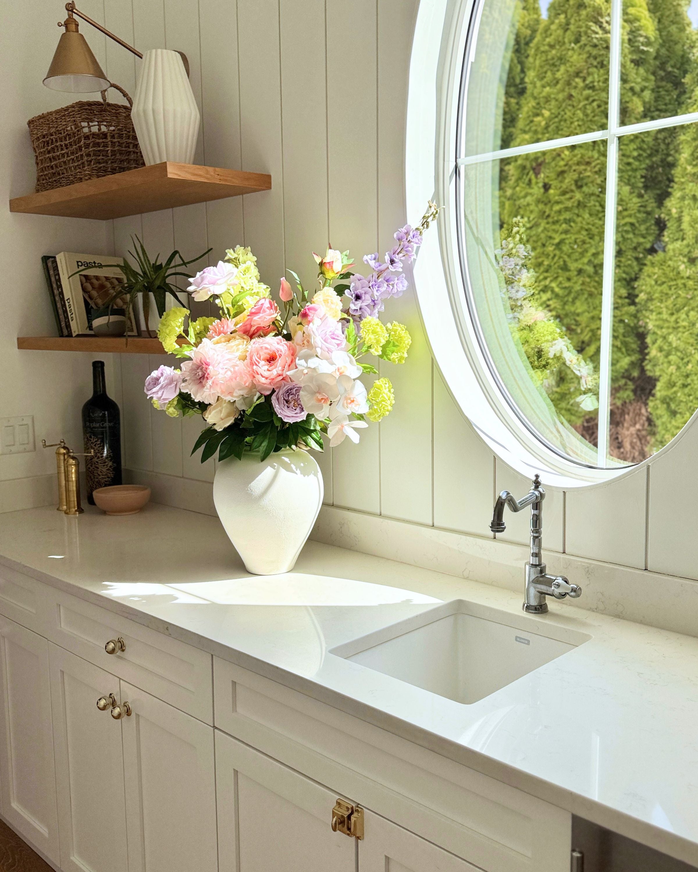 Kitchen counter with a vase of realistic artificial flowers, sink, and round window. Faux peonies, faux roses, faux hydrangea, and faux orchids in an artificial flower bouquet for My Faux Florist Canada.