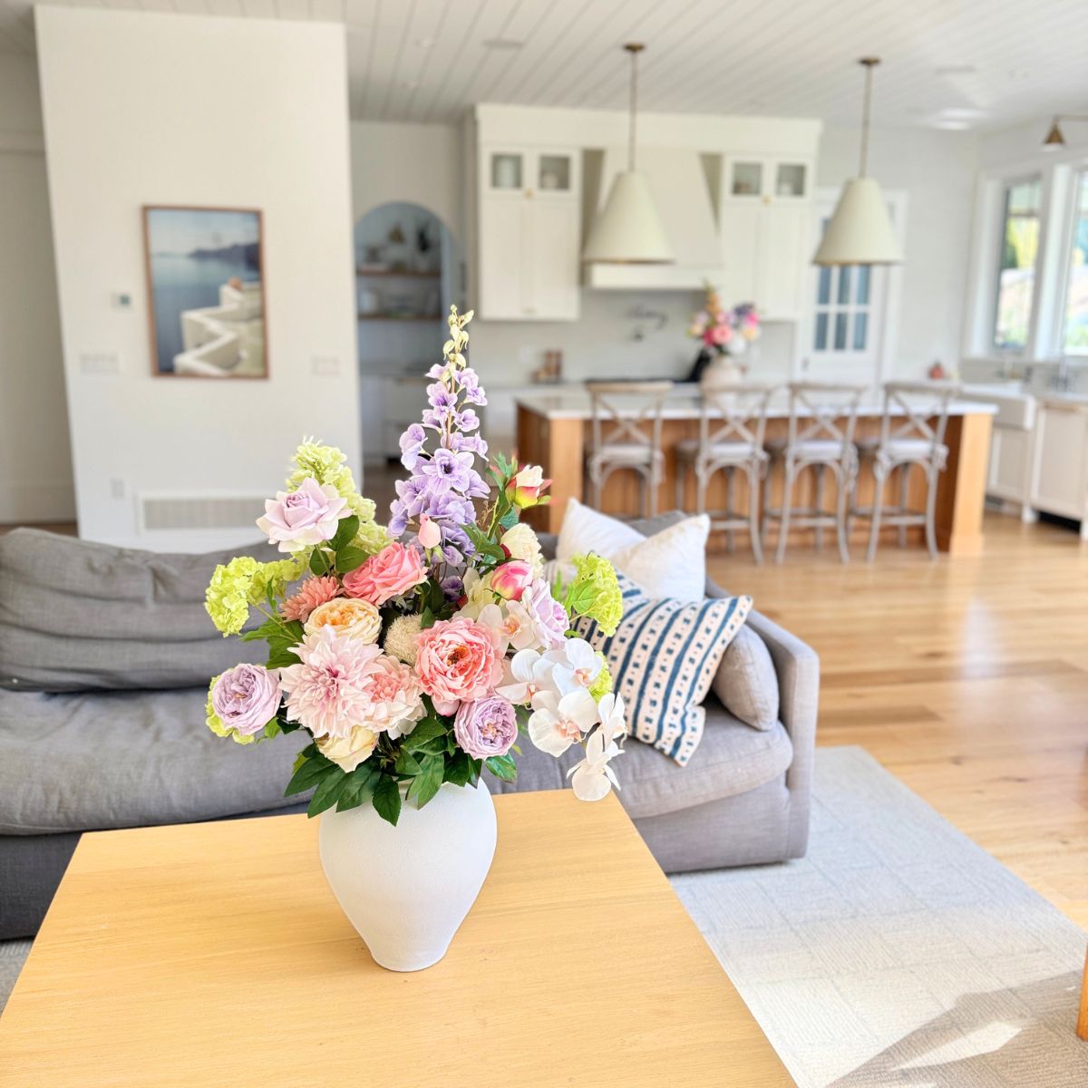 Floral arrangement of realistic artificial flowers in a white vase on a wooden table with a living room and kitchen in the background. Faux peonies, faux roses, faux hydrangea, and faux orchids in an artificial flower bouquet for My Faux Florist Canada.