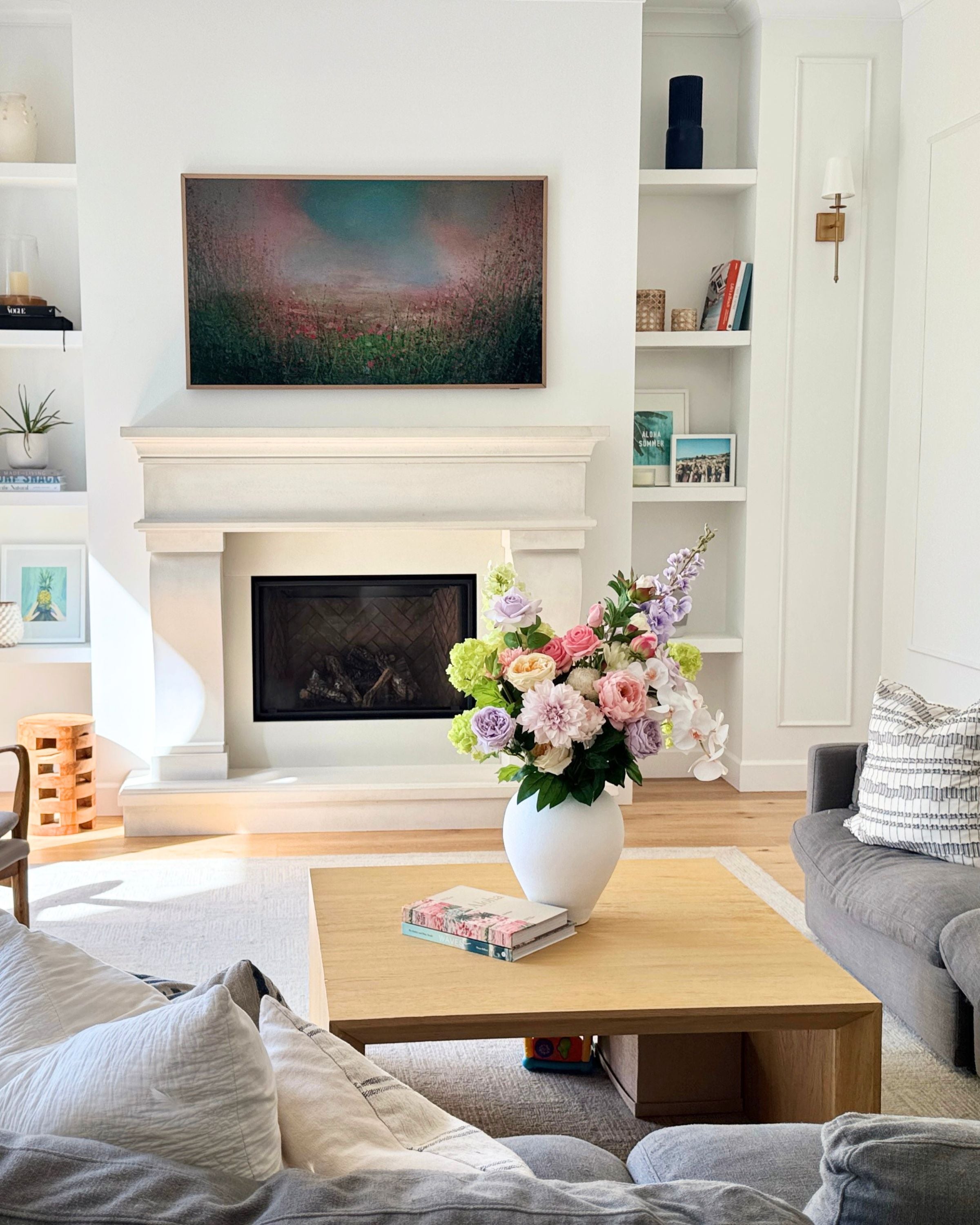 Living room with a fireplace, coffee table, and decorative items including a  large white vase of realistic artificial flowers. Faux peonies, faux roses, faux hydrangea, and faux orchids in an artificial flower bouquet for My Faux Florist Canada.