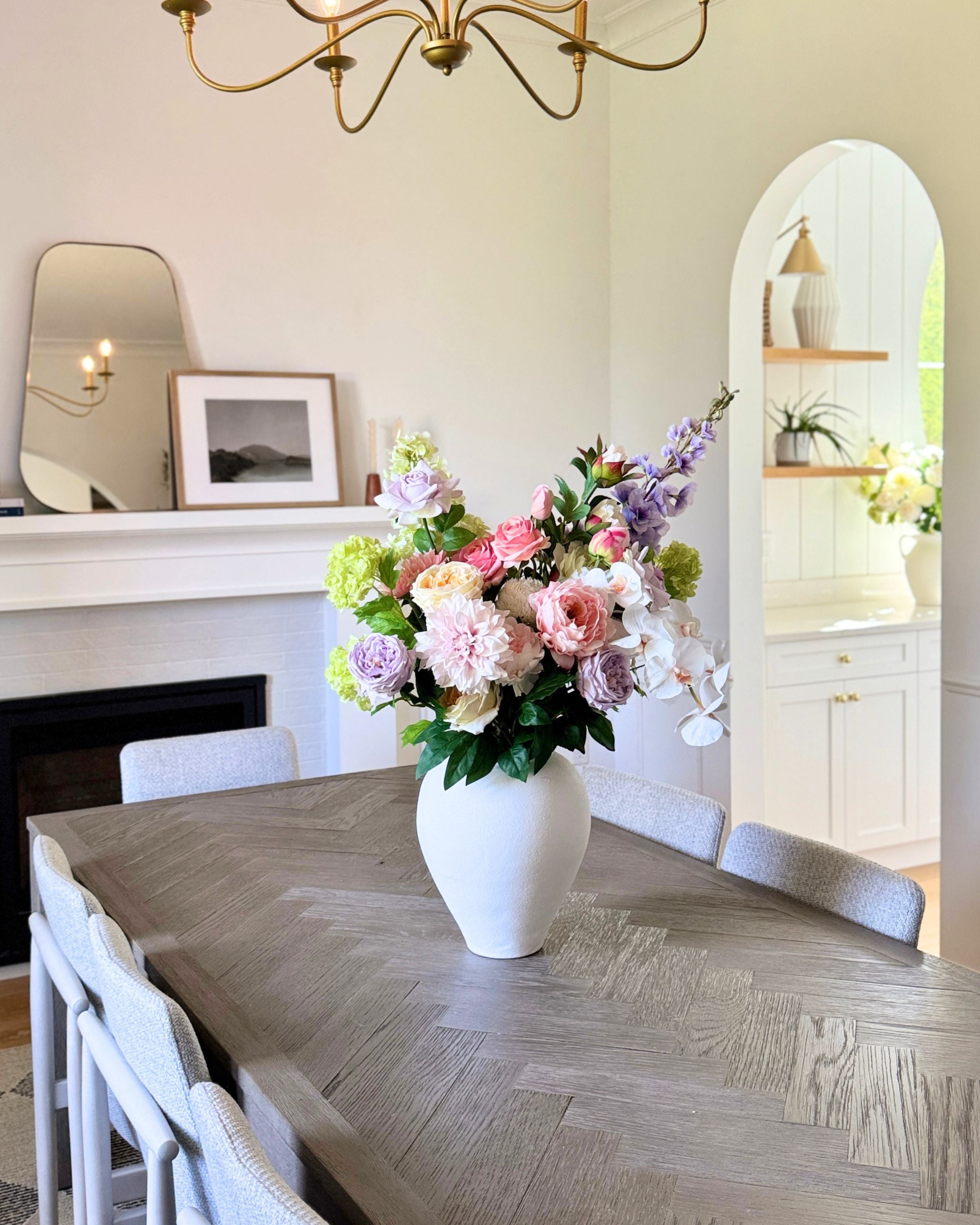 Floral arrangement of realistic artificial flowers in a white vase on a wooden dining table with a chandelier and mirrors in the background. Faux peonies, faux roses, faux hydrangea, and faux orchids in an artificial flower bouquet for My Faux Florist Canada.