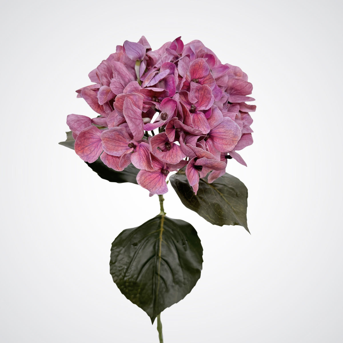 A single pink realistic artificial hydrangea stem with dark green leaves, isolated on a light gray background. From My Faux Florist Canada.