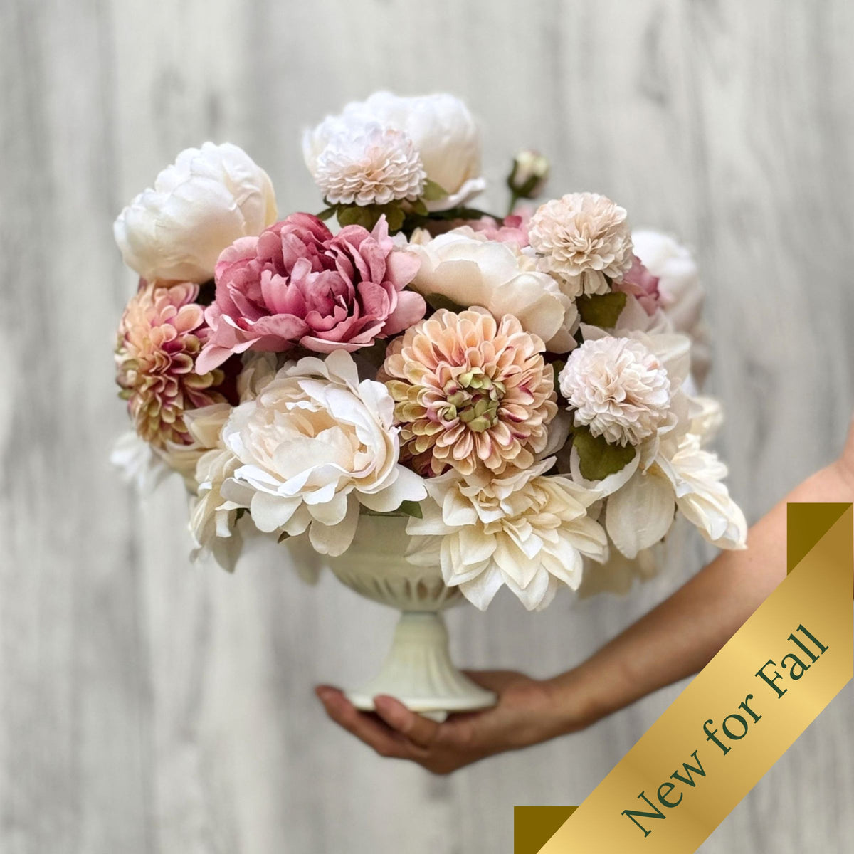 A large artificial flower arrangement for Fall in a white pedestal bowl, held by a person's hand against a light gray wood-textured background. The bouquet features cream, blush pink, peach, and mauve flowers with green leaves. Artificial flowers from My Faux Florist Canada.