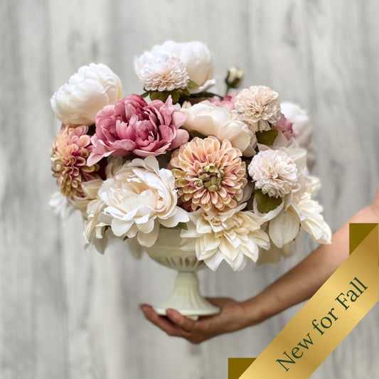 A large artificial flower arrangement for Fall in a white pedestal bowl, held by a person's hand against a light gray wood-textured background. The bouquet features cream, blush pink, peach, and mauve flowers with green leaves. Artificial flowers from My Faux Florist Canada.