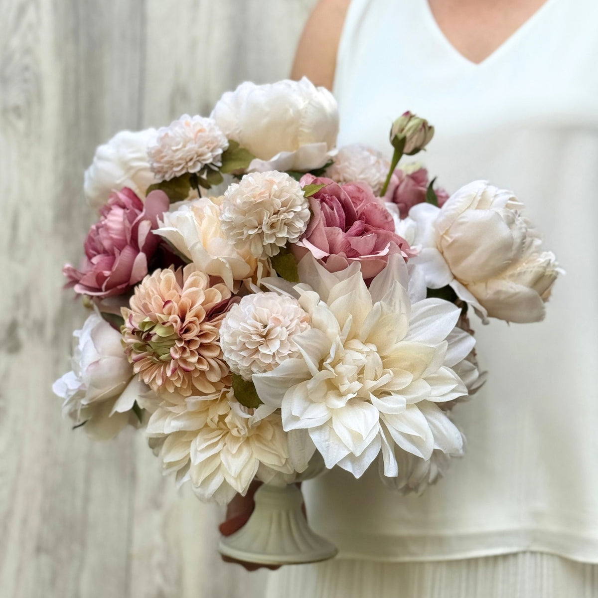 A close-up of a person holding a large bouquet of artificial flowers for Fall in a white sleeveless top. The bouquet features cream, blush pink, peach, and white peonies and dahlia-like blooms with a small white pedestal vase. Artificial flowers from My Faux Florist Canada.