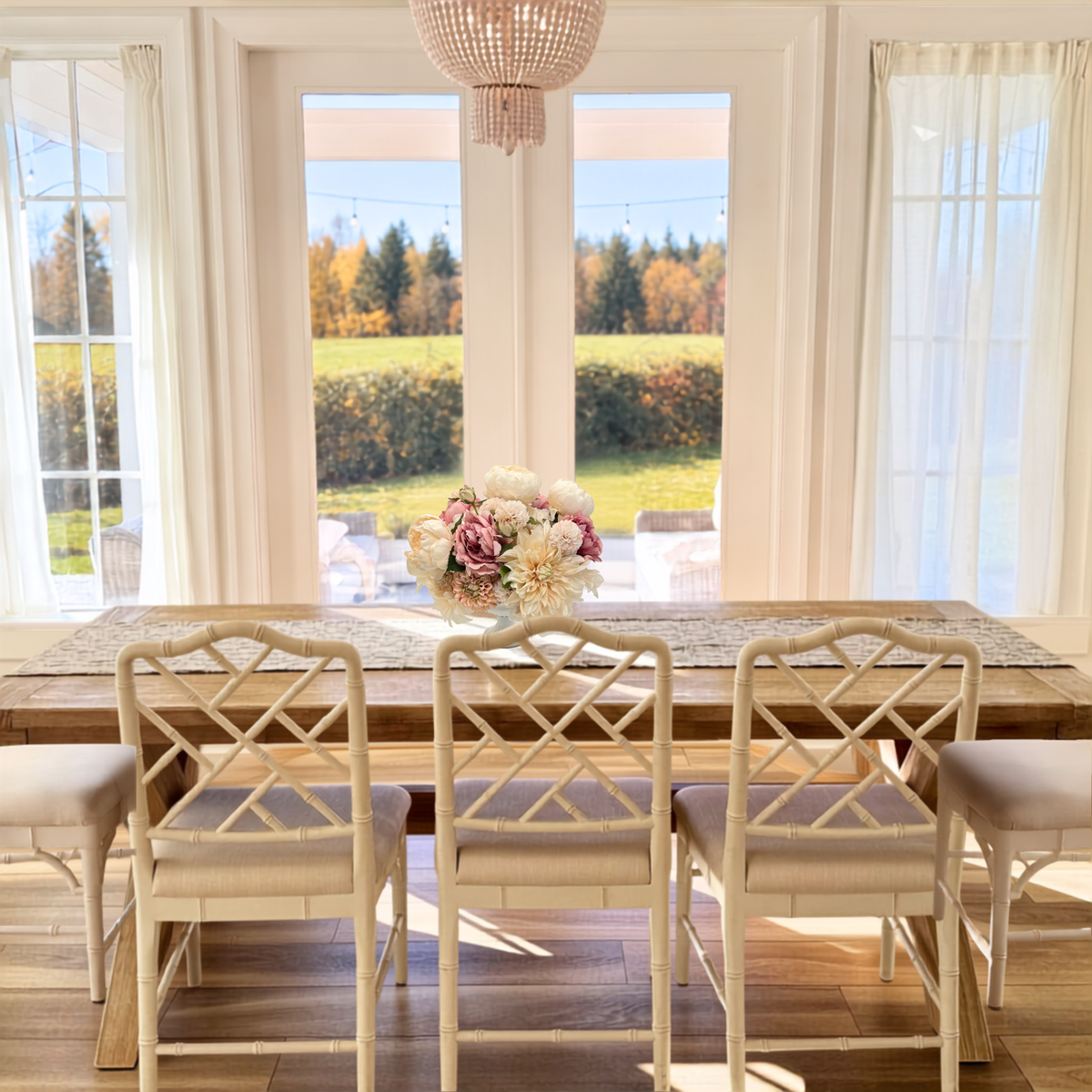 A dining room scene with a rectangular wooden table and four cream-colored lattice-back chairs with light beige upholstered seats. A floral centerpiece sits on the table - a faux flower arrangement for fall - and large windows behind it show a sunlit outdoor lawn and trees. Artificial flowers from My Faux Florist Canada.