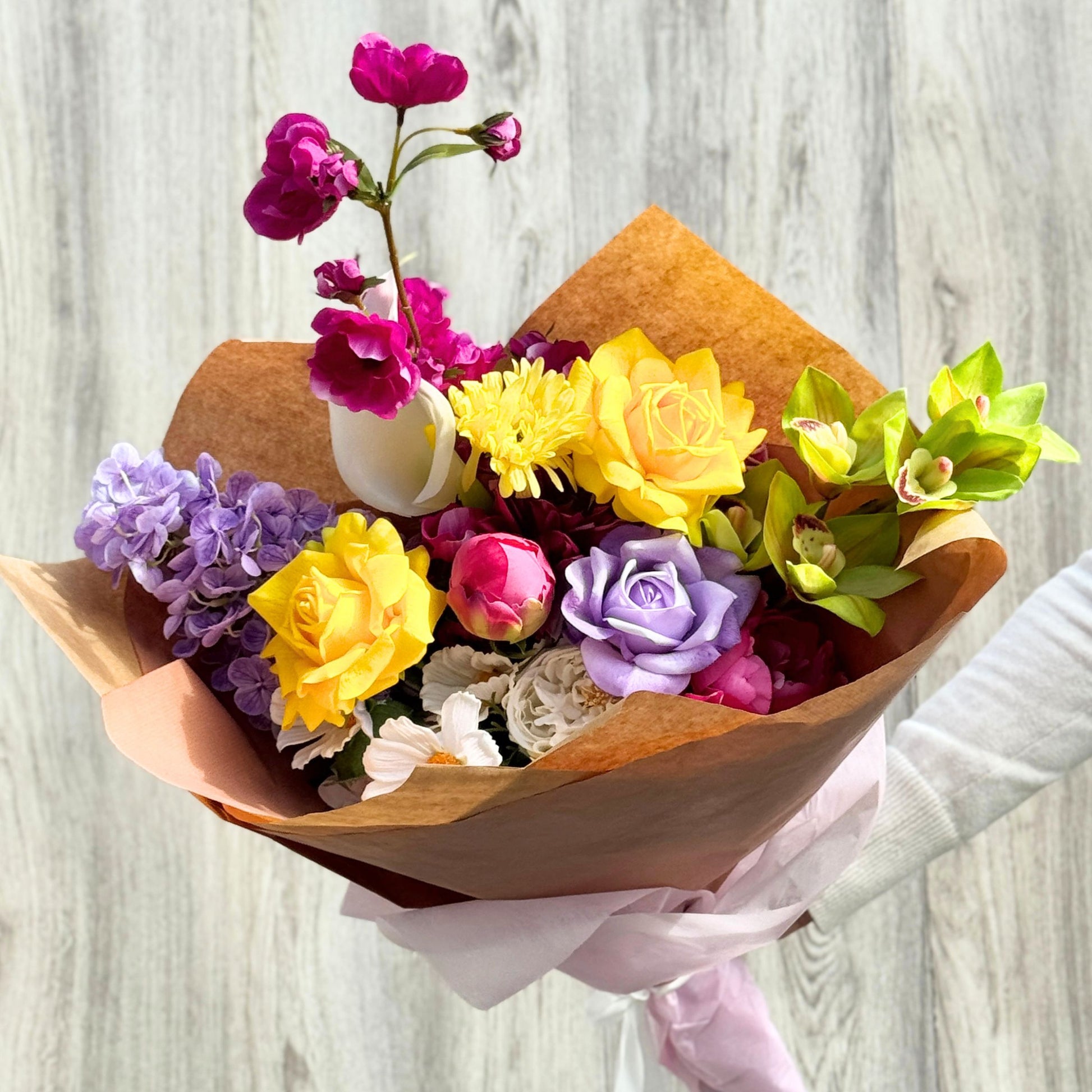 Bouquet of colorful realistic artificial flowers wrapped in brown paper on a light wooden background. Faux peonies, faux roses, faux hydrangea, and faux orchids in an artificial flower bouquet for My Faux Florist Canada.