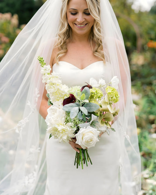 Bride standing outdoors holding an artificial wedding bouquet of realistic artificial flowers in, whites and greens from My Faux Florist Canada.